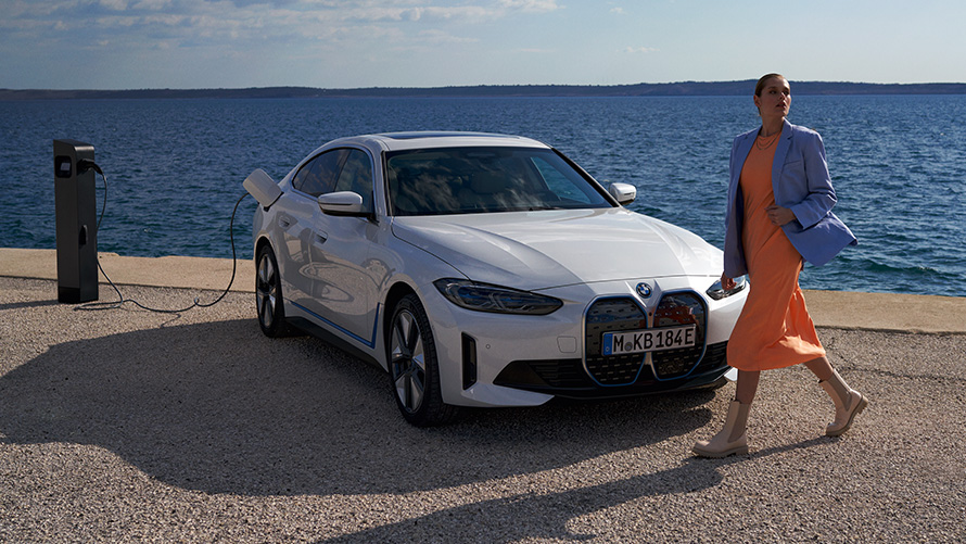Vista de tres cuartos del frontal BMW i4 eDrive40 G26 2021 en punto de carga Mujer delante del BMW i4 eDrive40 G26 2021 Mineralweiss, vista de tres cuartos del frontal, en un punto de carga, frente al mar