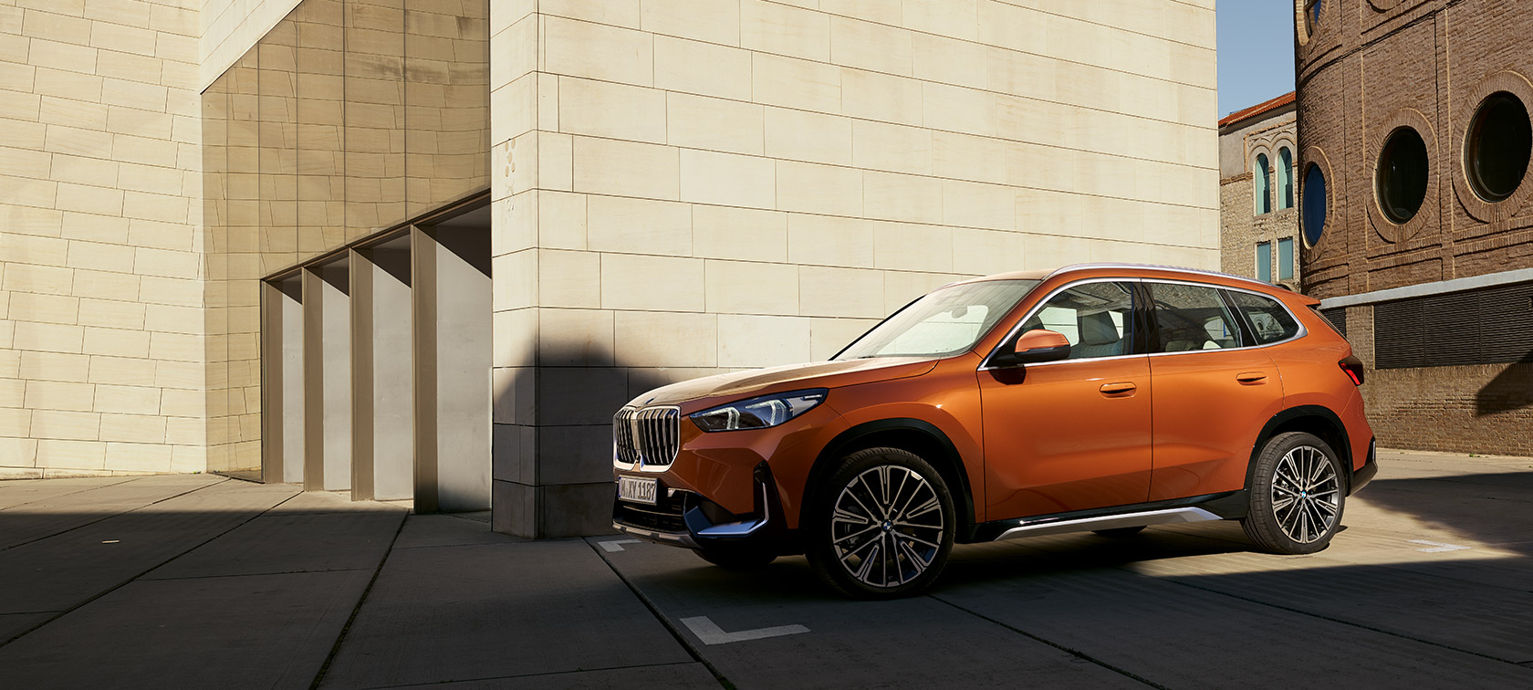 BMW U11 X1 xLine Utah Orange metalizado, vista de tres cuartos del frontal BMW U11 X1 xLine Utah Orange metalizado, vista de tres cuartos del frontal, parado delante de un edificio